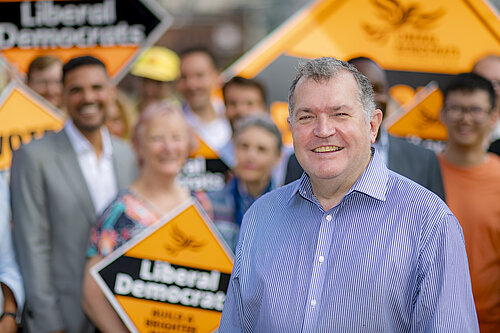 Headshot of Chris Maines with a blurred background consisting of activists holding up orange lib dem diamond placards
